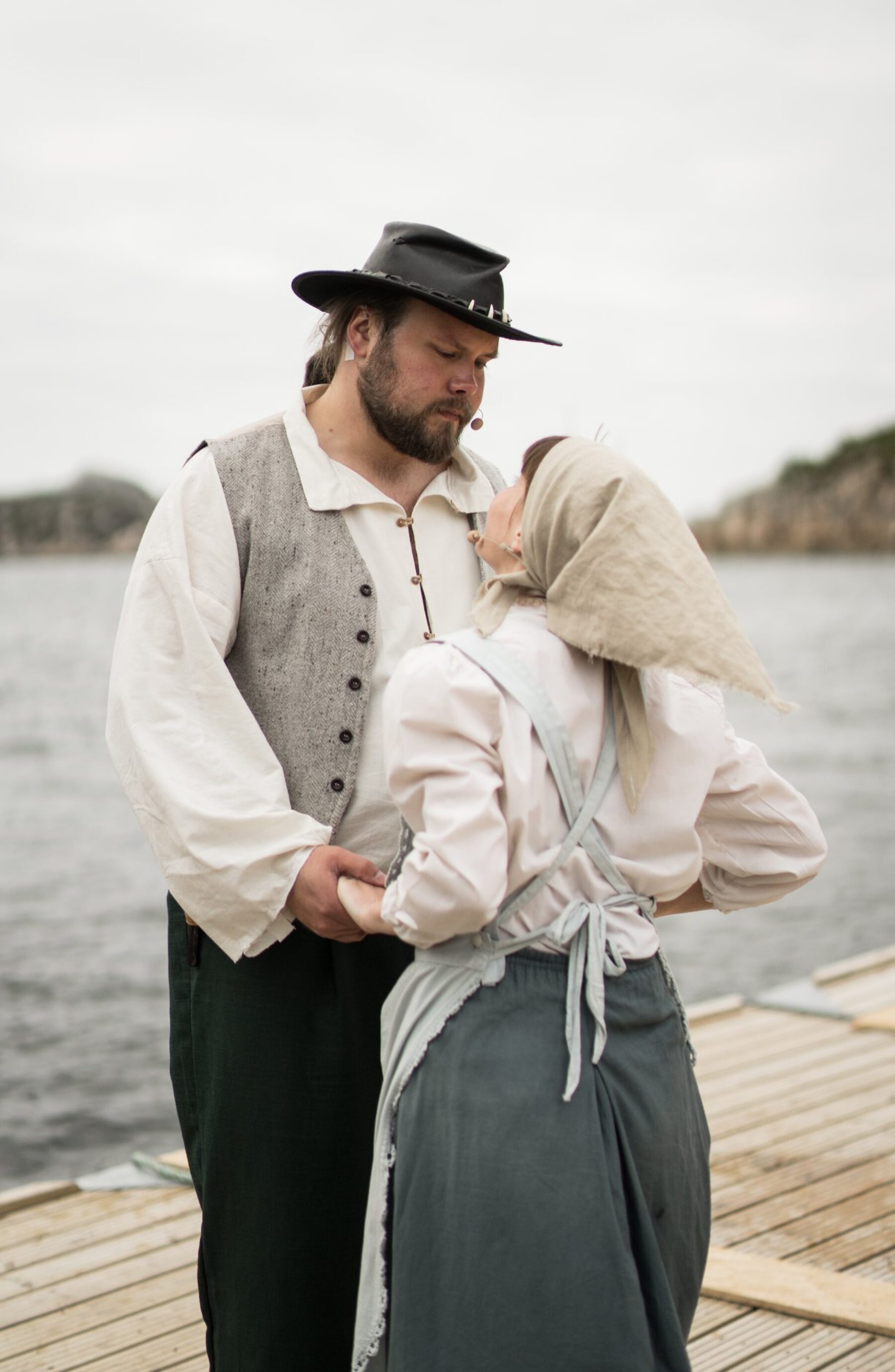 En skjeggete mann i hatt og grå vest holder hånden til en kvinne i skaut og forkle mens de ser på hverandre. Bak dem er fjorden.