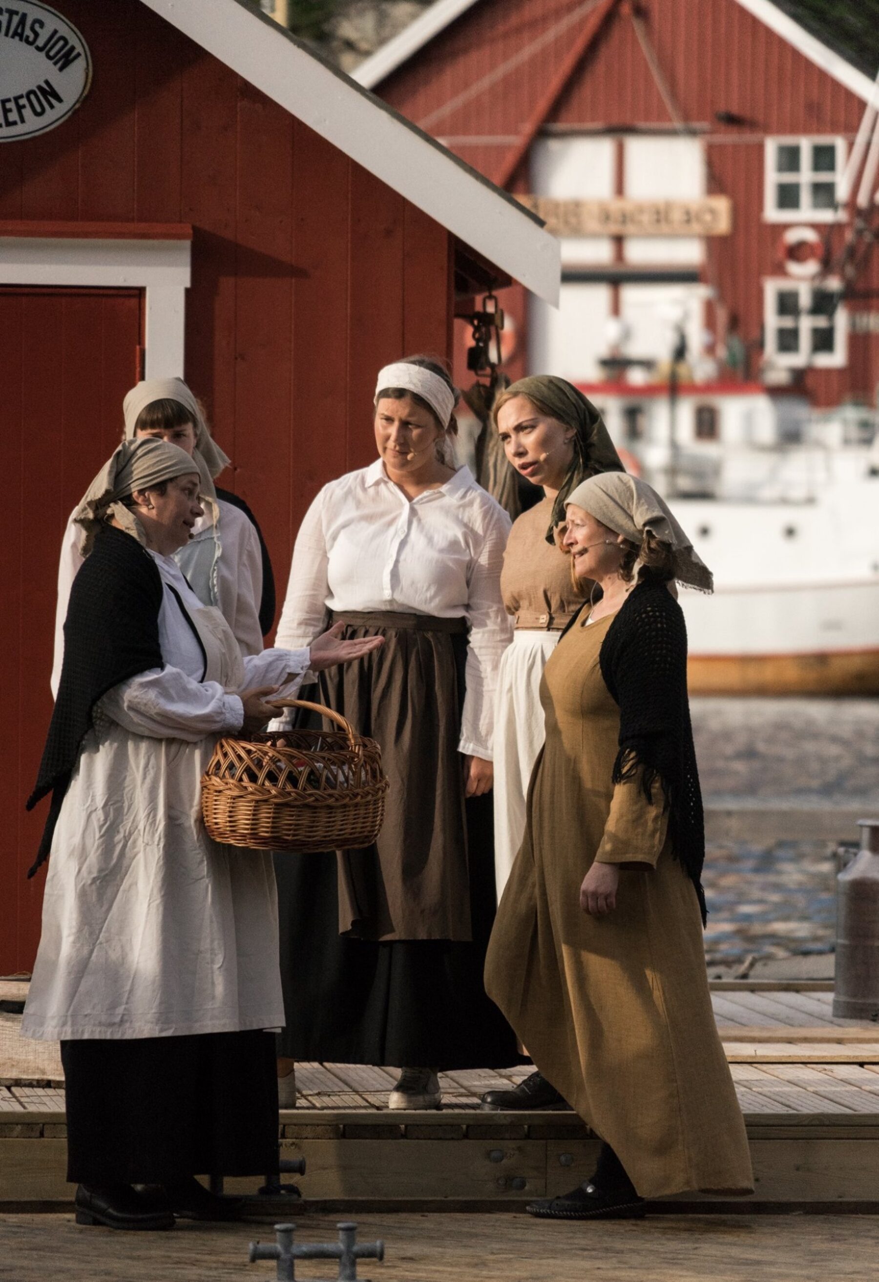 Kvinner i skaut og forklær står i samtale på brygga. Én holder en flettet kurv, og en rød sjøbu og en båt ses i bakgrunnen.