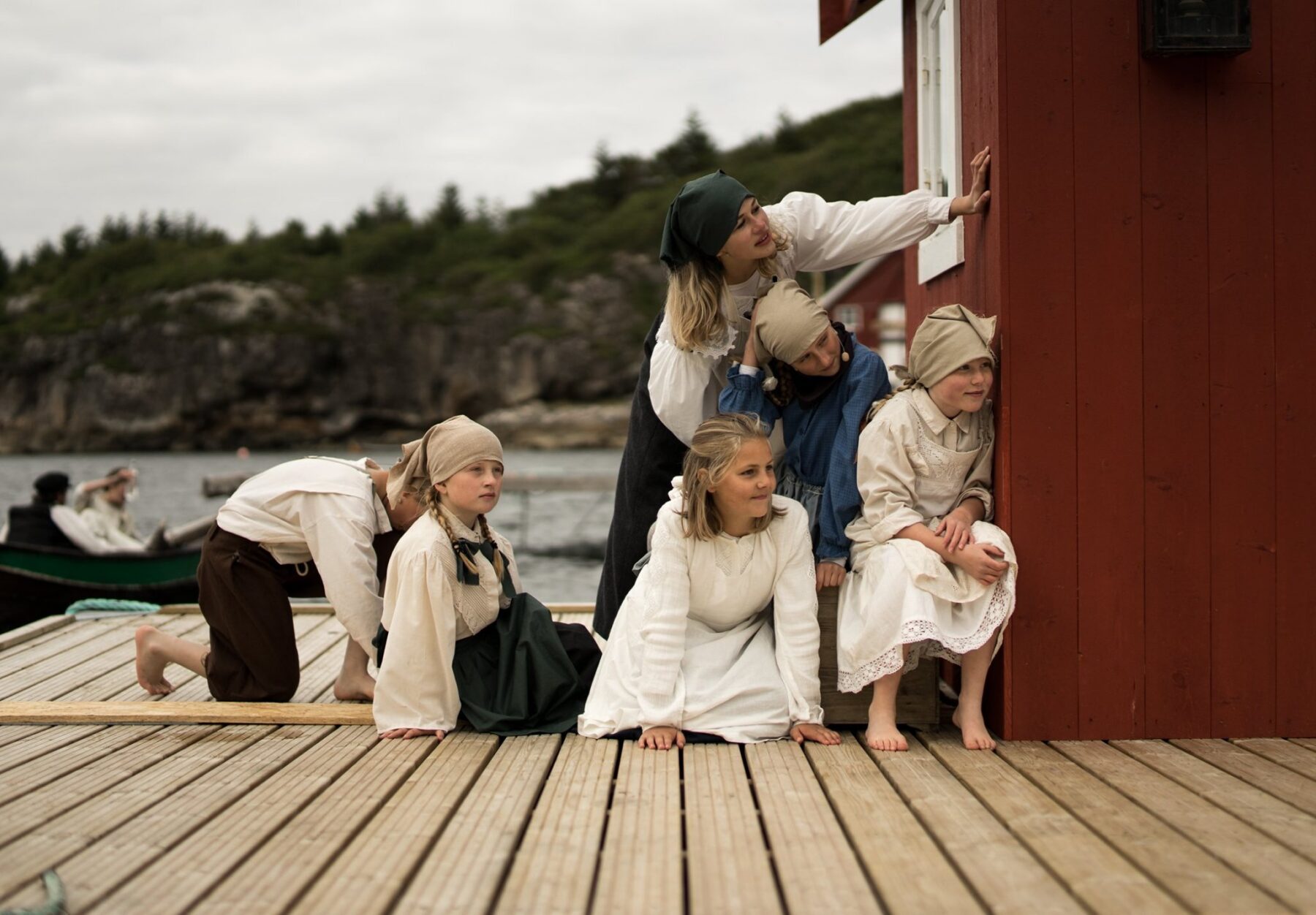 En gruppe barn i gammeldagse klær og skaut kryper og sitter på brygga og titter nysgjerrig rundt hjørnet på et rødt naust.