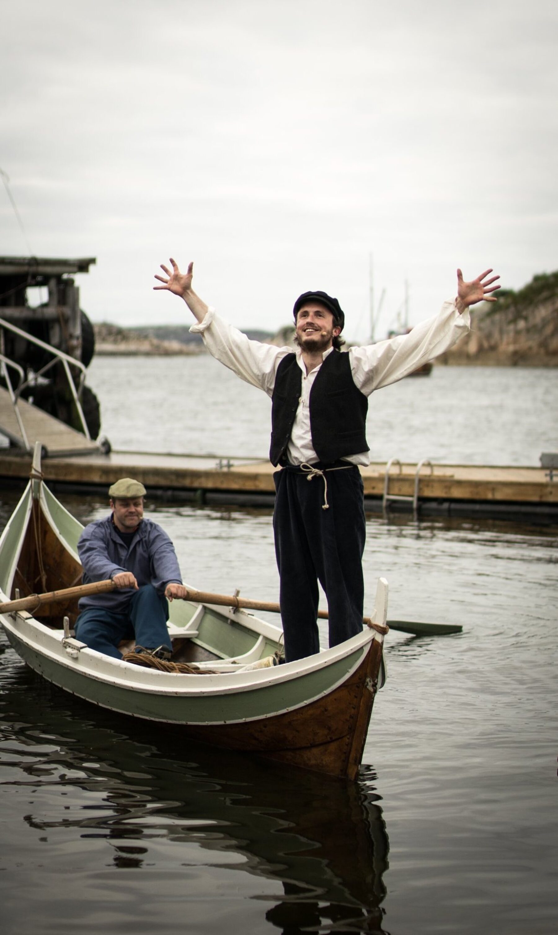 En ung mann med sixpence står i en robåt med armene løftet i jubel, mens en annen mann ror. Bak dem ligger brygga og fjorden.