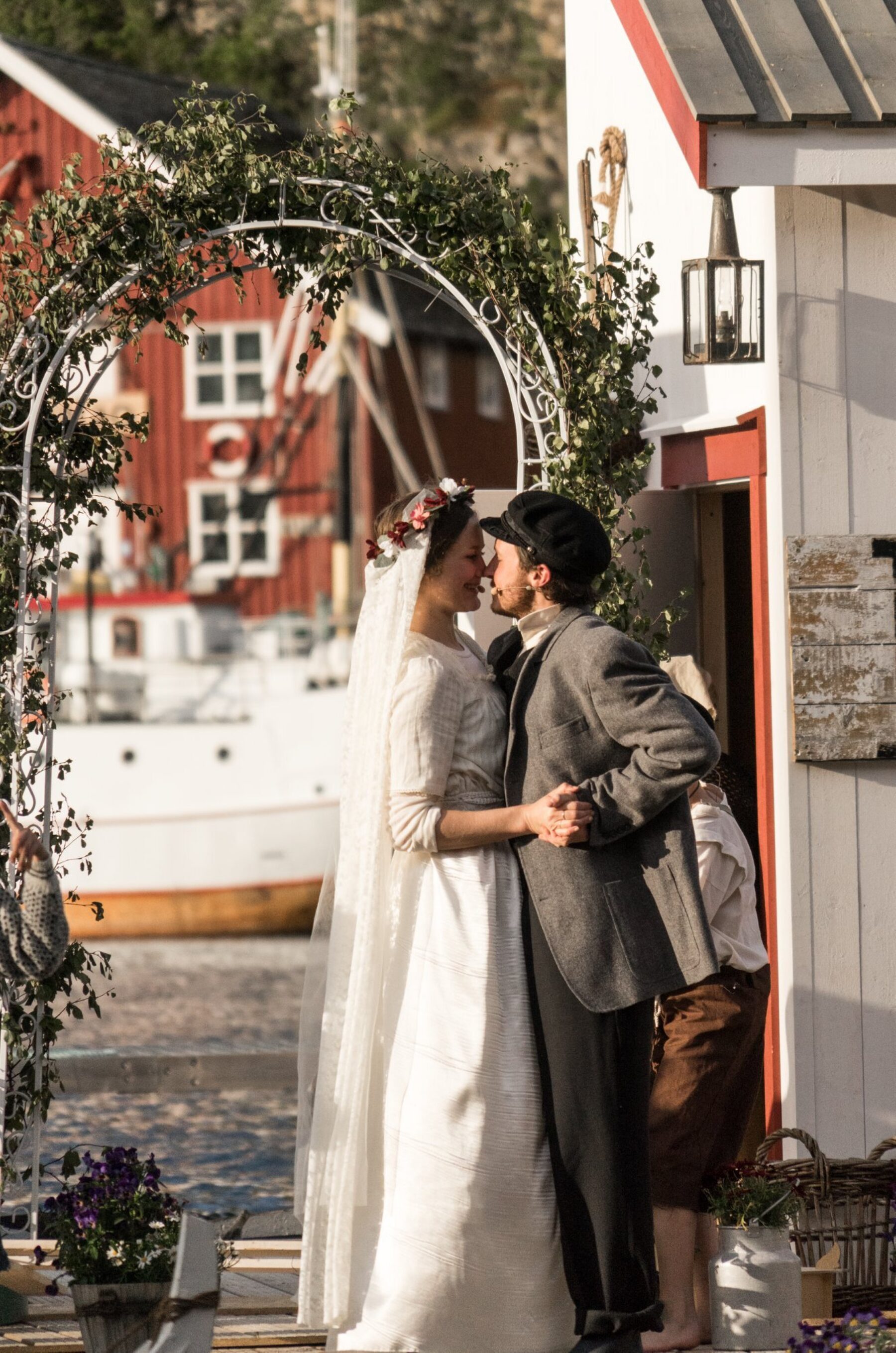 Et brudepar kysser under en blomsterbue på brygga i kveldssol. Bruden har slør og blomsterkrans, brudgommen sixpence og grå jakke.