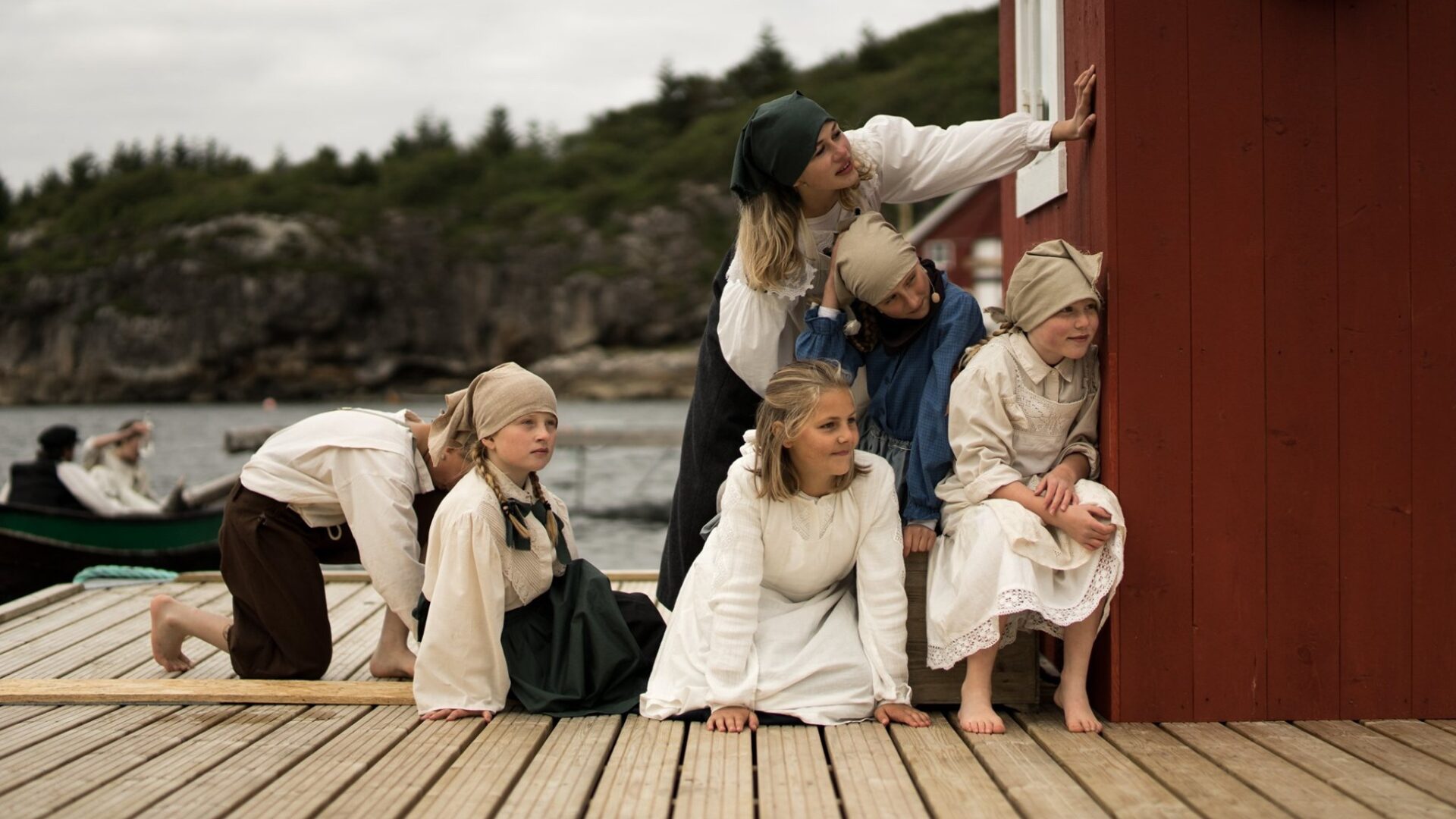 En gruppe barn i gammeldagse klær og skaut kryper og sitter på brygga og titter nysgjerrig rundt hjørnet på et rødt naust.