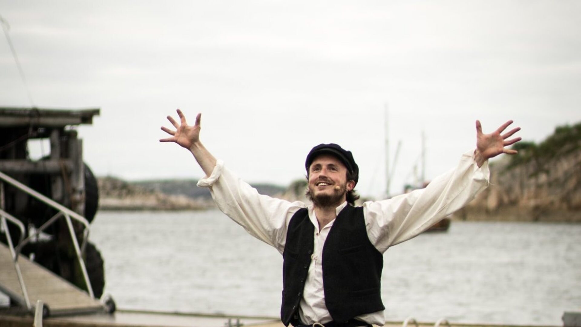 En ung mann med sixpence står i en robåt med armene løftet i jubel, mens en annen mann ror. Bak dem ligger brygga og fjorden.