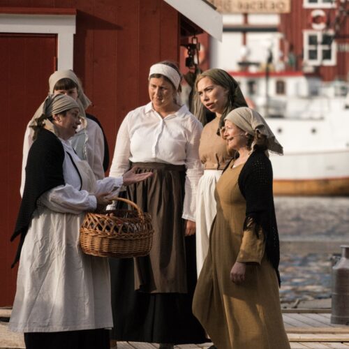 Kvinner i skaut og forklær står i samtale på brygga. Én holder en flettet kurv, og en rød sjøbu og en båt ses i bakgrunnen.
