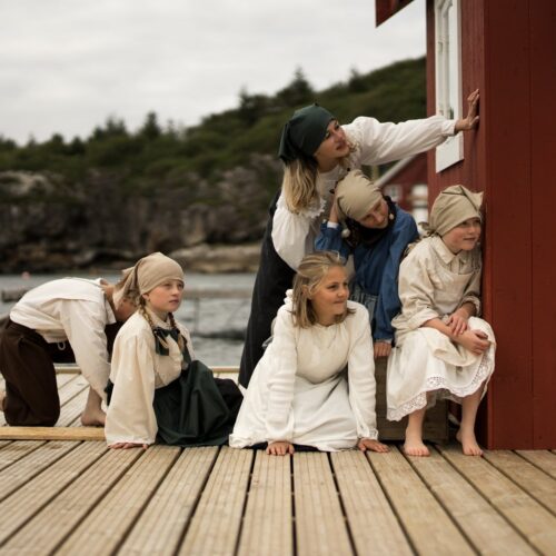 En gruppe barn i gammeldagse klær og skaut kryper og sitter på brygga og titter nysgjerrig rundt hjørnet på et rødt naust.