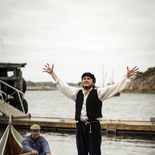 En ung mann med sixpence står i en robåt med armene løftet i jubel, mens en annen mann ror. Bak dem ligger brygga og fjorden.