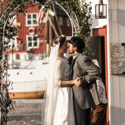Et brudepar kysser under en blomsterbue på brygga i kveldssol. Bruden har slør og blomsterkrans, brudgommen sixpence og grå jakke.
